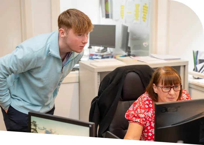 A TMQ employee in a light blue top leans over to look at a computer screen being used by another TMQ accountant in a red floral top sitting at a desk, discussing accountancy services in an office environment.