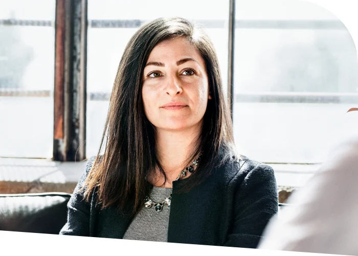 A woman with long, dark hair and a confident expression sits in a well-lit room, wearing a dark blazer and a statement necklace, embodying the essence of business coaching.
