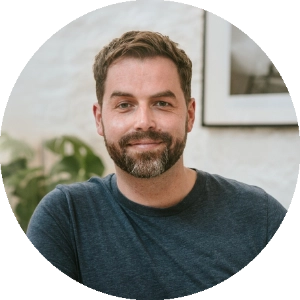 A person with short brown hair and a beard, wearing a blue shirt, sits indoors in front of a white wall with a framed picture, offering coaching advice.
