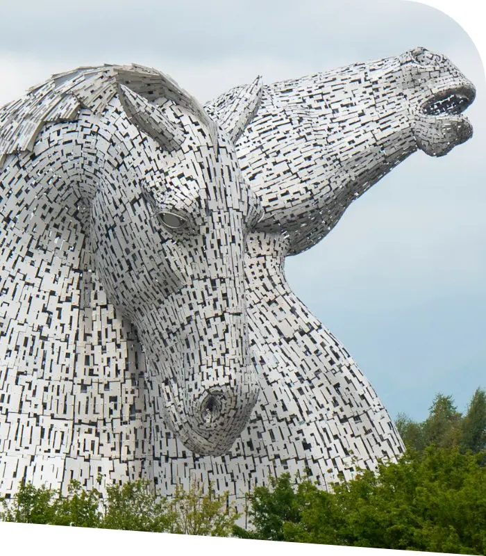 A close-up view of The Kelpies, two large horse-head sculptures made of steel, set against a cloudy sky with green foliage at the base, near the towns where Falkirk and Larbert accountants ply their trade.
