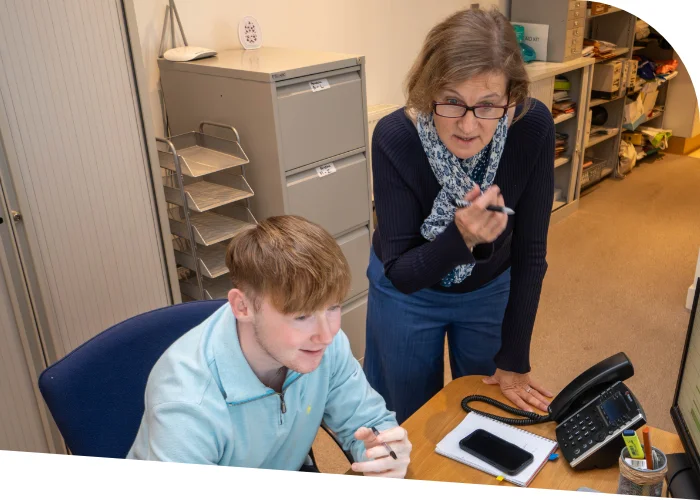 Susanne Robertson leans over and talks to an employee sitting at a desk, who is looking at a computer monitor. The desk, cluttered with office supplies, a phone, and a smartphone, hints at their discussion on effective business pricing strategies.