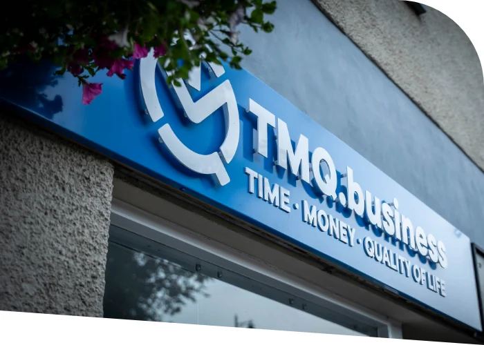 Close-up of a blue sign on a building that reads "TMQ.business - TIME • MONEY • QUALITY OF LIFE" with the TMQ Accountants & Business Advisers logo above. The surroundings show part of a wall and some greenery in Bridge of Allan.