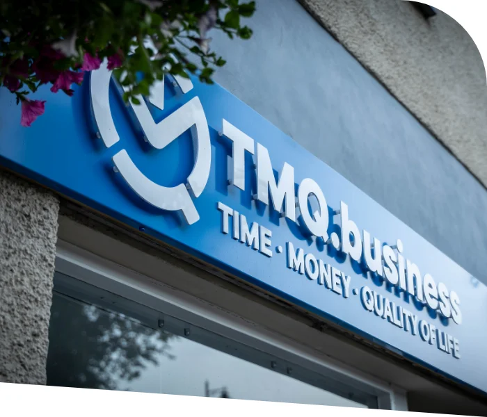 Close-up of a blue sign on a building that reads "TMQ.business - TIME • MONEY • QUALITY OF LIFE" with the TMQ Accountants & Business Advisers logo above. The surroundings show part of a wall and some greenery in Bridge of Allan.